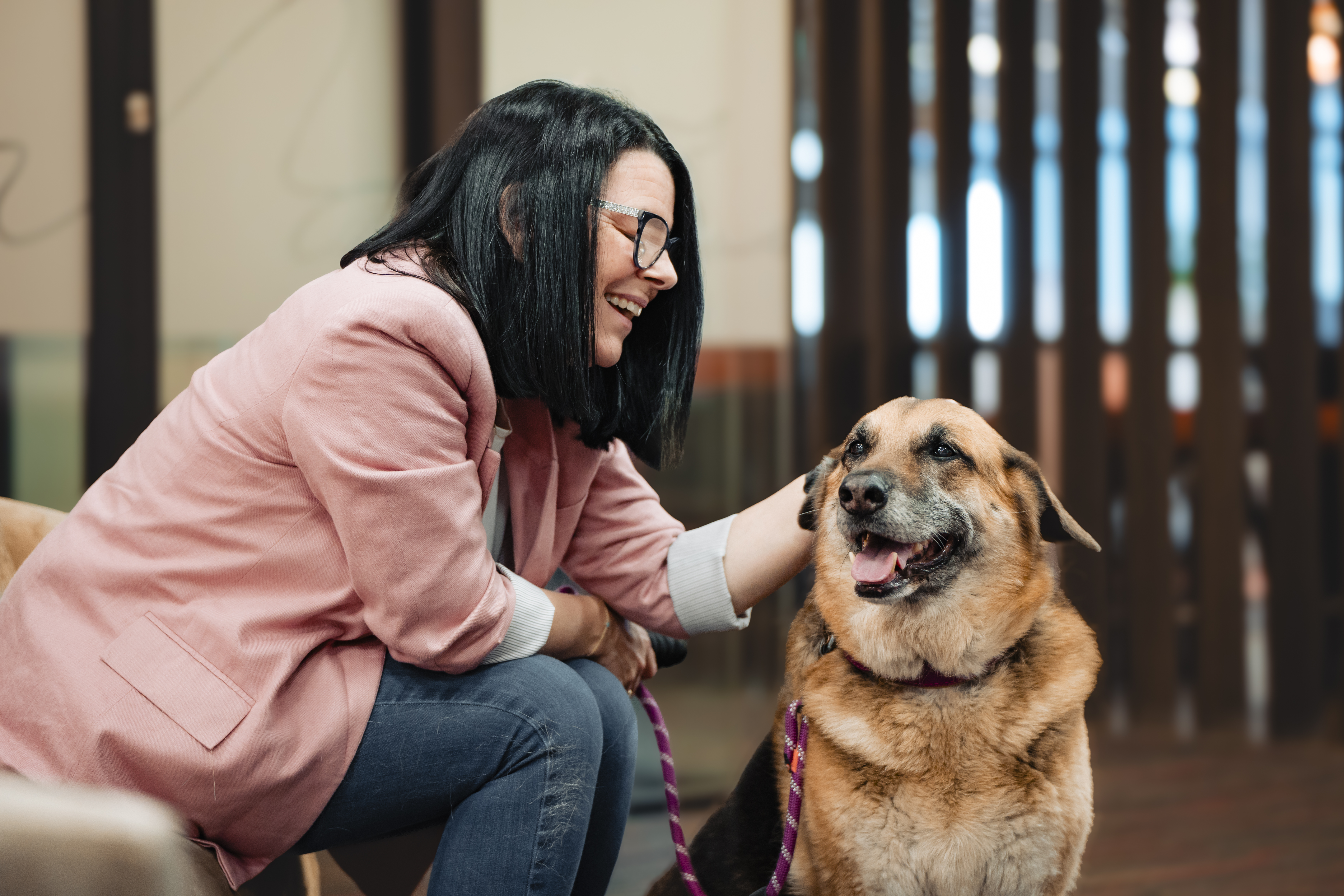 Femme en blazer rose souriant à un chien à l'intérieur.
